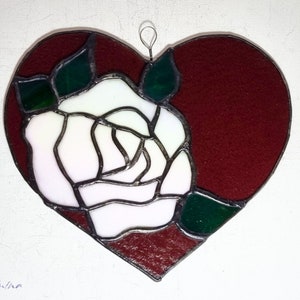 May include: A stained glass heart-shaped ornament with a white rose and green leaves on a burgundy background.