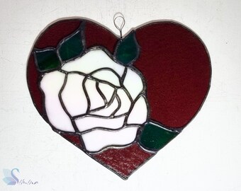 May include: A stained glass heart-shaped ornament with a white rose and green leaves on a burgundy background.
