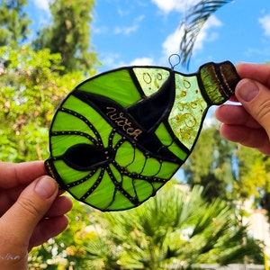 May include: A green glass bottle with a black witch hat and gold accents. The bottle is decorated with gold dots and the word "witch" is written on the hat.