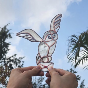 May include: A stained glass owl with a white and brown color scheme, holding a red envelope in its talons. The owl is in flight with its wings spread out.