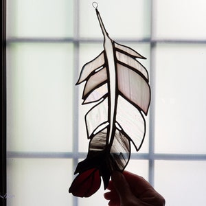 May include: A stained glass feather with a red, white, and black color scheme. The feather is hanging in a window.