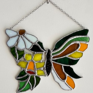 May include: Stained glass butterfly suncatcher with a daisy design. The butterfly features a mix of colors including white, green, yellow, orange, brown, and black. It hangs from a silver chain, perfect for adding a touch of nature-inspired art to any space.