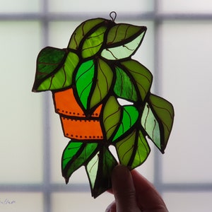 May include: A stained glass hanging of a potted plant with green leaves and an orange pot. The leaves are outlined in black and the pot has a brown and white pattern.