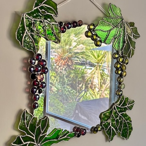 May include: A decorative mirror with a stained glass grape vine design. The frame features green leaves and clusters of purple and green grapes. The mirror is rectangular and hangs from a white string.