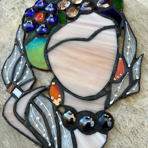 May include: A stained glass artwork depicting a stylized female figure with a heart-shaped face. The figure is adorned with a crown of flowers and a necklace of beads. The artwork is made with various colors of glass, including blue, green, red, and gold.