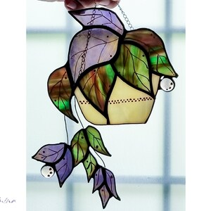 May include: A stained glass hanging featuring a potted plant with purple, green, and brown leaves. The pot is yellow with brown dots and the leaves have small clear glass beads hanging from them.