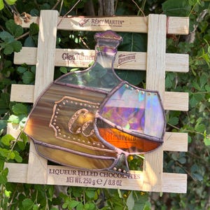 May include: Stained glass art piece depicting a liquor bottle and a glass of Amaretto. The artwork is framed by a wooden lattice and includes the text "LIQUEUR FILLED CHOCOLATES NET WT. 250 g / 8.8 OZ". The piece has brown, orange, and purple hues.