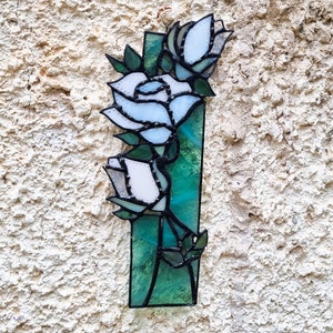 May include: A stained glass window hanging on a white wall, featuring a bouquet of white roses with green leaves and stems. The roses are made of stained glass with a blue and white color scheme.