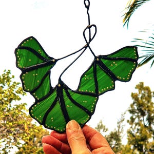 May include: A green stained glass ginkgo leaf hanging ornament with gold accents. The leaf is suspended by a wire and has a delicate, intricate design.