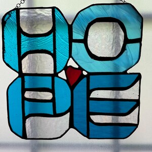 May include: Stained glass art piece spelling out the word "HOPE" in a stylized design. The letters are composed of blue, silver, and red glass, outlined in black. A chain is attached for hanging.