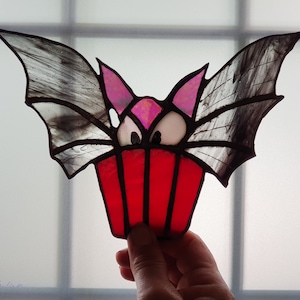 May include: A stained glass bat with pink ears and eyes, holding a red cupcake.