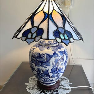 May include: A blue and white porcelain lamp base with a stained glass lampshade featuring a floral design. The lampshade is made of clear and blue glass and has a white interior.