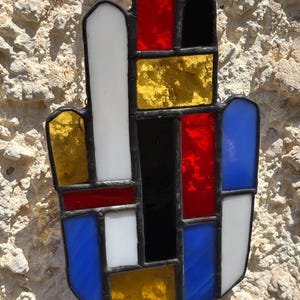 May include: A stained glass hand-shaped suncatcher with a black border. The design features rectangular and arched shapes in red, yellow, black, white, and blue. It hangs from a silver chain.