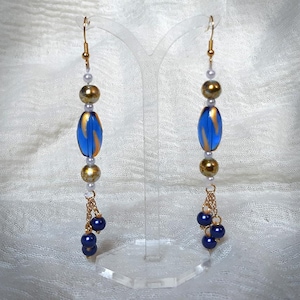 May include: A pair of dangle earrings with gold-colored hooks. Each earring features a combination of gold, blue, and white beads, including a blue oval bead with gold accents. Small navy blue beads hang from a gold chain.
