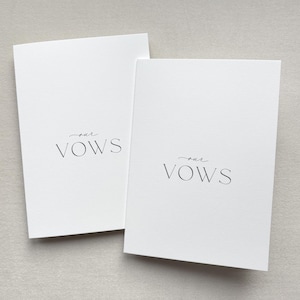 Op de afbeelding: Twee witte geloftenboeken met de woorden "our vows" gedrukt in een sierlijk, cursief lettertype. De rechthoekige boeken zijn lichtjes schuin geplaatst tegen een neutrale achtergrond, passend bij een bruiloft of ceremonie.