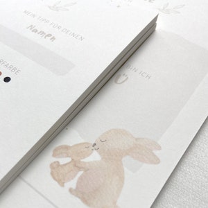 May include: Two watercolor illustrations of bunnies hugging. The bunnies are light brown and beige. The text "Bin Ich" is written in cursive on the right side of the image.
