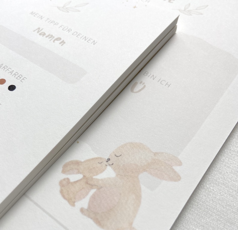 May include: Two watercolor illustrations of bunnies hugging. The bunnies are light brown and beige. The text "Bin Ich" is written in cursive on the right side of the image.