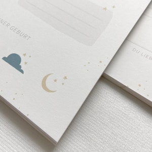 May include: A white card with a blue cloud and a gold crescent moon with stars. The text "FINER GEBURT" is printed on the card.
