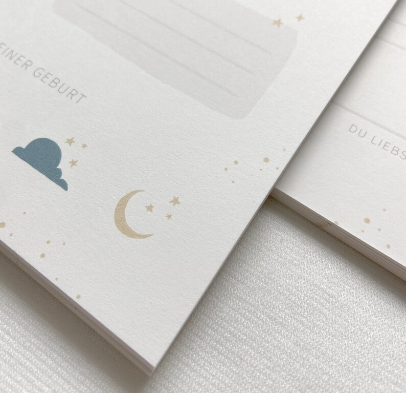 May include: A white card with a blue cloud and a gold crescent moon with stars. The text "FINER GEBURT" is printed on the card.