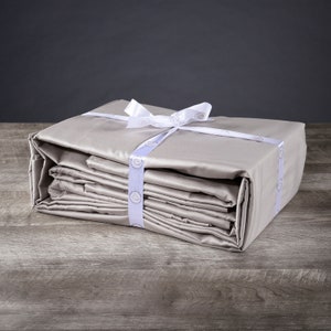 May include: A stack of beige bed sheets tied with a white ribbon. The sheets are folded neatly and have a subtle sheen.