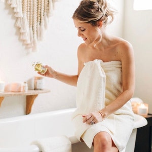 May include: A woman wrapped in a cream-colored towel sits on the edge of a white bathtub, holding a small glass bottle. The bottle contains a clear liquid. A wooden shelf with candles and a decorative wall hanging are in the background.