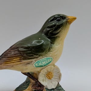 May include: A ceramic bird figurine with a green and tan body, perched on a branch with a white flower. The bird has a yellow beak and is marked with a "Beswick England" sticker. The figurine is approximately 6 inches long.