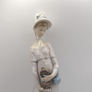 May include: A porcelain figurine of a woman in a long, pale purple dress and hat, holding a small brown dog and a blue purse. The figurine is standing next to a sculpted bush. The overall theme is a Victorian era lady.