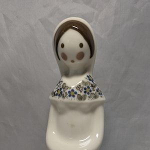 Puede incluir: Una figurita de cerámica blanca de una mujer con un sombrero marrón y un vestido con estampado floral azul y blanco. La figurita tiene una franja azul alrededor de la parte inferior del vestido.