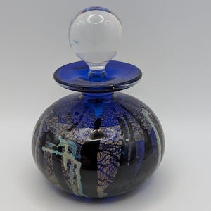 May include: A round, blue glass perfume bottle with a clear glass stopper. The bottle has a dark blue base with a decorative pattern of black, silver, and teal. The stopper is a clear, round shape.
