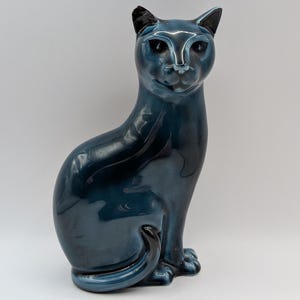 May include: A teal ceramic cat ornament in a seated pose. The cat has black detailing on its ears and eyes, with a glossy finish. The figurine is a dark teal colour and is set against a plain white backdrop.