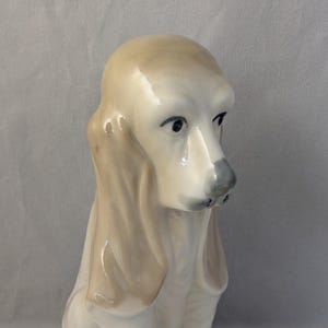 May include: A ceramic figurine of a seated dog with long, floppy ears. The dog is primarily white with tan ears and accents, and black spots on its paws. The dog has a sad expression.
