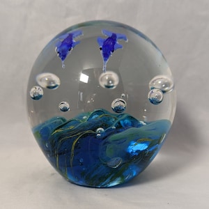 Puede incluir: Un pisapapeles de vidrio con dos delfines azules nadando en un océano azul y verde con burbujas.