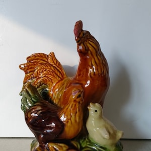 May include: A ceramic rooster figurine with a brown, yellow and green glaze. The rooster is standing with its wings spread and its head tilted back. A smaller rooster and a chick are nestled beside it.