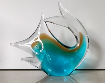 May include: A glass sculpture of a blue and green fish with a clear tail fin. The fish is in a curled position, with its tail curled around its body.