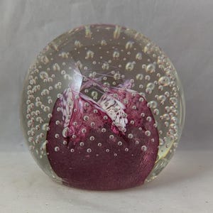 Può includere: Un fermacarte di vetro trasparente con un vortice viola e bianco all'interno. Il fermacarte è pieno di piccole bolle d'aria.