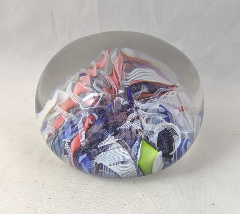 May include: A spherical glass paperweight featuring an internal design of swirling ribbons in shades of pink, blue, white, and black, with a single solid lime green accent piece encased within the clear, polished glass orb.