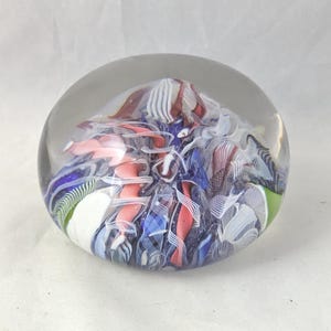 May include: A clear glass paperweight with a rounded dome shape containing an internal design of swirling ribbons in shades of blue, pink, white, and dark brown. The interior features intricate patterns of layered glass filaments and opaque segments.