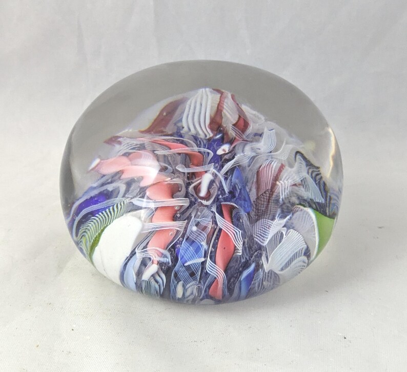 May include: A clear glass paperweight with a rounded dome shape containing an internal design of swirling ribbons in shades of blue, pink, white, and dark brown. The interior features intricate patterns of layered glass filaments and opaque segments.