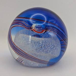 May include: A clear glass paperweight with a blue, red, and white swirl design. The paperweight has a rounded shape with a frosted, cloud-like interior. The blue glass has a glossy finish.