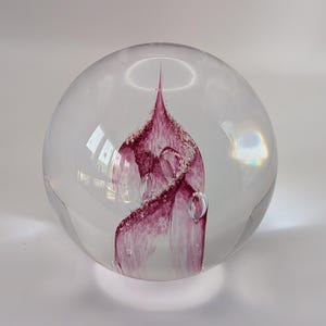 May include: A clear glass paperweight with a swirling, abstract design in shades of purple and white. The design has a textured, frosted appearance and is encased within the spherical glass. The paperweight has a smooth, polished surface.