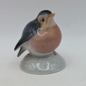 May include: A ceramic bird ornament in shades of grey, black, and pink. The bird has a pink breast and grey wings, with a black and grey head. It is perched on a light grey base, a decorative item.