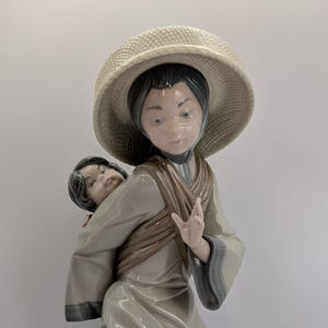 May include: A porcelain figurine of a woman carrying a child. The woman wears a large hat and a long dress in neutral colours. The child is carried in a sling. The figurine has a calm expression. The figurine is made of porcelain.