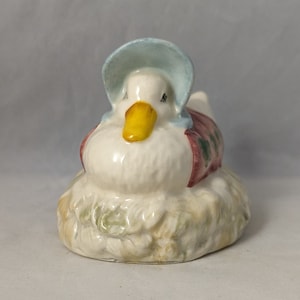 May include: A white ceramic figurine of a duck wearing a blue bonnet and a red and green dress. The duck is sitting on a bed of green and brown leaves.