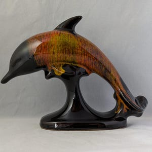 Puede incluir: Una figurita de delfín de cerámica con una base negra y un esmalte multicolor en tonos naranja, amarillo y marrón.