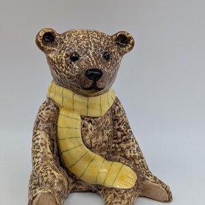 May include: A ceramic bear figurine with a textured brown finish, featuring a glossy yellow scarf with thin green stripes wrapped around its neck. The bear has dark, round eyes and a black nose, sitting in an upright position with a smooth surface.