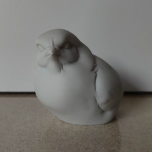 Puede incluir: Una figurita de pájaro de cerámica blanca con un cuerpo redondeado y una cara detallada. El pájaro tiene un pequeño pico y los ojos cerrados.