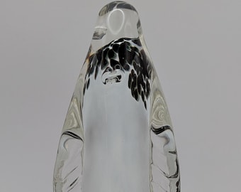 May include: A clear glass figurine shaped like a penguin, featuring a smooth, rounded body with internal black and white speckled patterns concentrated near the head and neck area, and defined wing-like indentations on the sides.