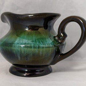 May include: A green and brown glazed ceramic creamer with a flared lip and a handle. The creamer has a glossy finish and a unique swirl pattern.