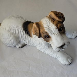 May include: A white porcelain figurine of a brown and white terrier dog lying down. The dog has brown ears and a brown patch on its back.
