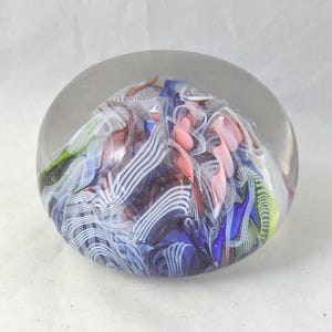 May include: A clear glass paperweight with a rounded dome shape containing an internal design of swirling ribbons in shades of blue, white, pink, and green. The interior features intricate patterns of opaque and translucent glass filaments.